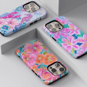 May include: Three colorful phone cases with floral and sea creature designs. One case features a lobster and seahorse, another has pink flowers, and the third showcases orange and pink blooms. Each case has a black border and a camera cutout.