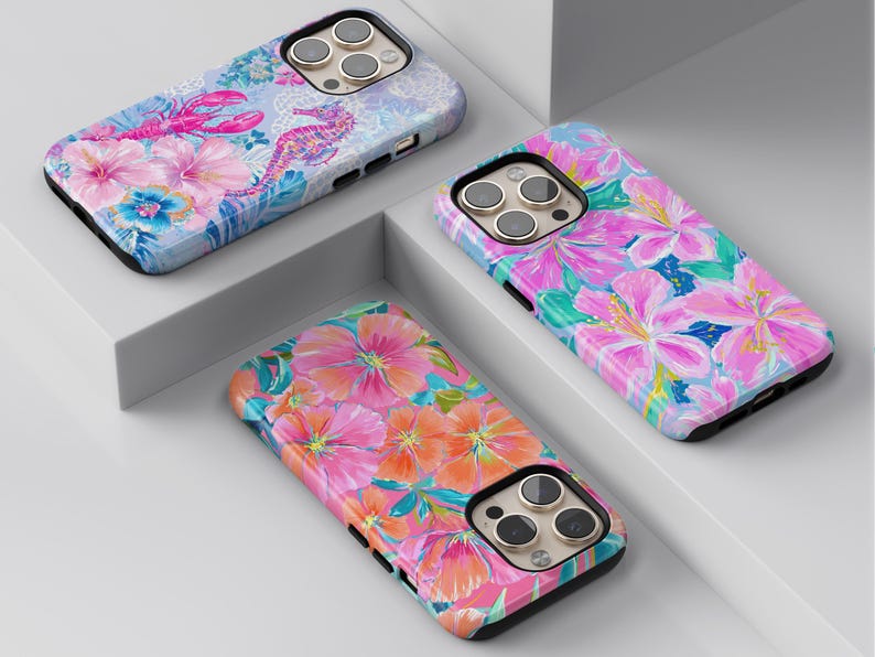 May include: Three colorful phone cases with floral and sea creature designs. One case features a lobster and seahorse, another has pink flowers, and the third showcases orange and pink blooms. Each case has a black border and a camera cutout.