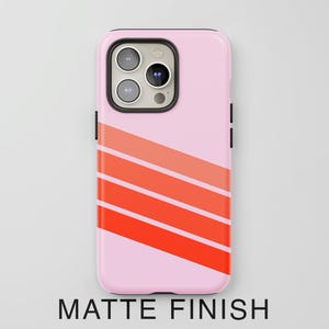 May include: A light pink smartphone case with a matte finish featuring three diagonal stripes in varying shades of orange and red across the lower portion. The case has a black camera module cutout with three circular lenses and a flash.