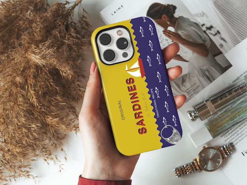 May include: A yellow phone case with a blue and white sardine can design. The can says "Original Sardines In Olive Oil Traditional Recipe".