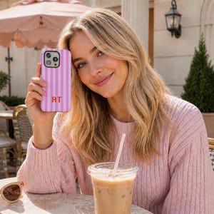 May include: A pink and white vertically striped smartphone case featuring the letters RT in a bold orange font. The case has a cutout for a triple-lens camera system and is designed to fit a modern smartphone with a sleek, protective exterior.