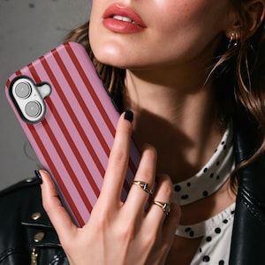 May include: A pink and red striped phone case. The case is held in front of a person. The phone case has a camera cutout and buttons. The person is wearing a black leather jacket and gold rings.