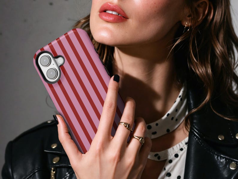May include: A pink and red striped phone case. The case is held in front of a person. The phone case has a camera cutout and buttons. The person is wearing a black leather jacket and gold rings.