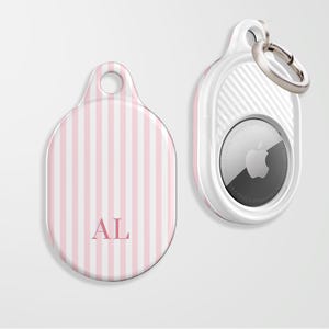 May include: Two AirTag holders, one with a pink and white striped pattern and the initials "AL". The other is white with a silver key ring. The AirTag itself is visible in the holder.