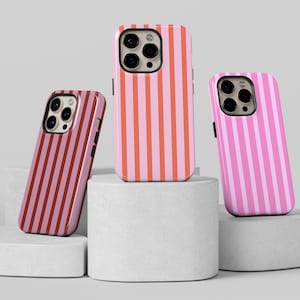 May include: Three phone cases with vertical stripe patterns in shades of pink and red. The cases are displayed on white cylindrical pedestals against a neutral background. The cases have camera cutouts.