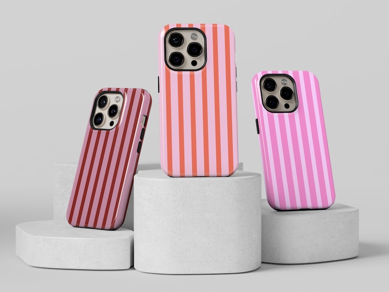 May include: Three phone cases with vertical stripe patterns in shades of pink and red. The cases are displayed on white cylindrical pedestals against a neutral background. The cases have camera cutouts.