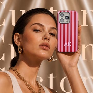 May include: A smartphone case featuring vertical stripes in alternating shades of dark pink and light pink. The case includes a triple-lens camera cutout at the top and the white capital letters AK printed in the center of the lower half of the case.