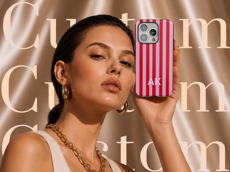 May include: A smartphone case featuring vertical stripes in alternating shades of dark pink and light pink. The case includes a triple-lens camera cutout at the top and the white capital letters AK printed in the center of the lower half of the case.