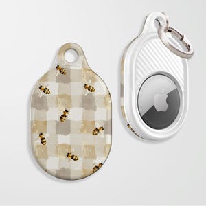 Puede incluir: Dos llaveros. Uno es un soporte blanco para Apple AirTag con un anillo plateado. El otro es una etiqueta decorativa con un patrón de cuadros beige y blanco e ilustraciones de abejas. La etiqueta de abeja tiene un agujero para sujetar.
