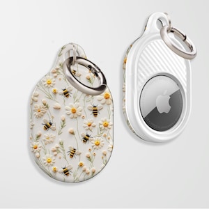 May include: White AirTag case with a floral pattern featuring white daisies and yellow bees. The case has a silver ring for attaching keys or a keychain.