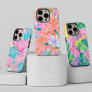 May include: Three phone cases with colorful floral and tropical leaf designs. The cases feature a variety of patterns, including pink and blue seashells, orange and pink flowers, and green and purple leaves. The cases are displayed on white pedestals.