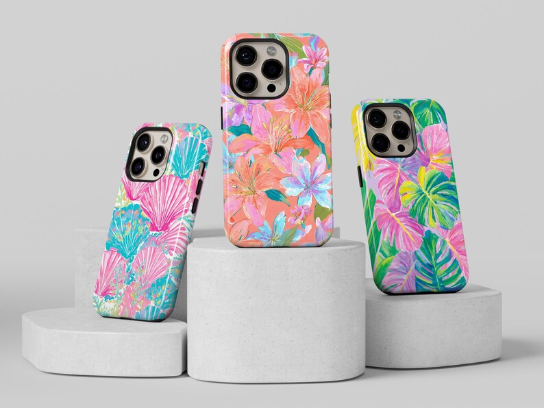 May include: Three phone cases with colorful floral and tropical leaf designs. The cases feature a variety of patterns, including pink and blue seashells, orange and pink flowers, and green and purple leaves. The cases are displayed on white pedestals.