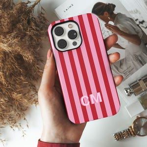 May include: A rectangular smartphone case featuring a vertical striped pattern in alternating shades of bright red and light pink. The letters CM are printed in a light pink sans-serif font in the lower center of the case, which has a cutout.