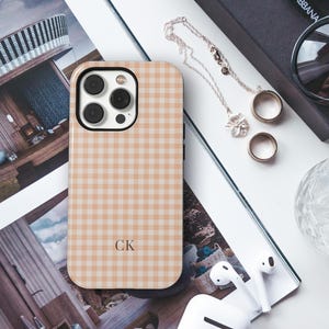 Puede incluir: Una funda rectangular para smartphone con un patr&oacute;n de cuadros vichy en color tostado claro y blanco. La funda tiene un recorte negro para la lente de la c&aacute;mara y las letras CK impresas en una fuente oscura en la parte inferior central.