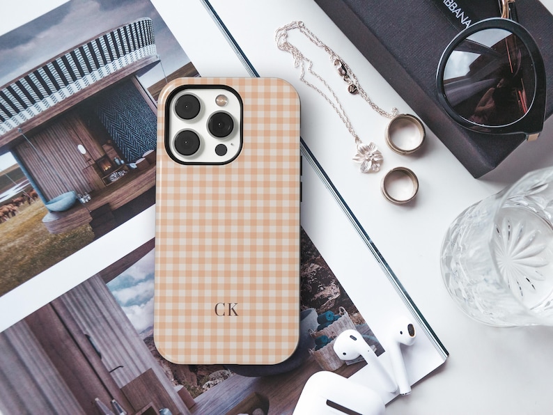 Puede incluir: Una funda rectangular para smartphone con un patr&oacute;n de cuadros vichy en color tostado claro y blanco. La funda tiene un recorte negro para la lente de la c&aacute;mara y las letras CK impresas en una fuente oscura en la parte inferior central.