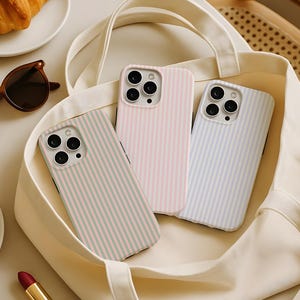 Puede incluir: Tres fundas para teléfono con patrones de rayas verticales en colores pastel: rosa, azul y verde. Las fundas están dentro de un bolso tote color crema. También se ven gafas de sol y cruasanes.