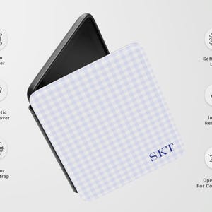 May include: A phone case with a light blue and white gingham pattern. The case has a black interior and features the initials "SKT" in navy blue. Additional features include vegan leather, a magnetic front cover, and an interior hand strap.