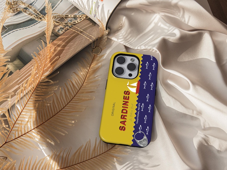 May include: A yellow and blue phone case with a sardine can design. The case features the text "ORIGINAL SARDINES" and a traditional recipe label. The case has a blue background with white fish silhouettes.