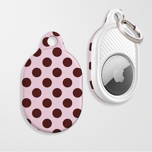 Könnte beinhalten: Eine rosa AirTag-Hülle mit braunem Polka-Dot-Muster, neben einem weiß-rosa AirTag-Halter mit einem silbernen Schlüsselring. Der AirTag hat das Apple-Logo.