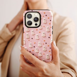 May include: A pink phone case featuring a pattern of white ghosts and orange stars. The case is designed for a smartphone with a triple lens camera.