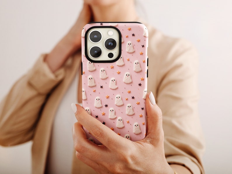 May include: A pink phone case featuring a pattern of white ghosts and orange stars. The case is designed for a smartphone with a triple lens camera.
