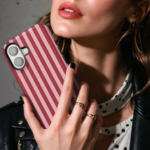 May include: A pink and red striped phone case with a camera lens cutout. The case is being held by a person's hand.