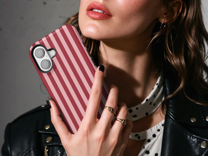 May include: A pink and red striped phone case with a camera lens cutout. The case is being held by a person's hand.