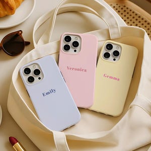 Puede incluir: Tres fundas de teléfono de colores pastel en un bolso tote crema. Las fundas son lavanda, rosa y amarillo, cada una con un nombre: Emily, Veronica y Gemma. Gafas de sol y cruasanes en el fondo.