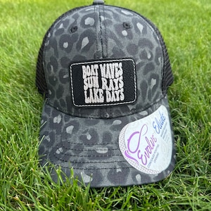 May include: Black and white leopard print trucker hat with a black leather patch that says "Boat Waves Sun Rays Lake Days". The hat has a black mesh back and a white logo on the side that says "Evolve & Eluude".