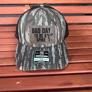 May include: Camouflage trucker hat with a brown leather patch that reads "BAD DAY TO BE A SPIKE" with a deer graphic. The hat has a black mesh back and an adjustable strap. The Richardson brand logo is visible.