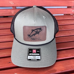 May include: A green and black trucker hat with a brown leather patch featuring a fish design. The hat has the text "EST. 1970 RICHARDSON. ORIGINAL 112 TRUCKER ADJUSTABLE" on the front.