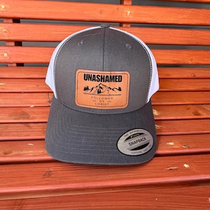 May include: A gray and white trucker hat with a curved brim. The hat features a brown leather patch with the word "UNASHAMED" and the phrase "FOLLOWER OF CHRIST". The hat has a snapback closure.