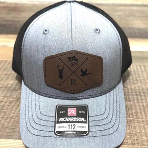 May include: A gray and black trucker hat with a brown leather patch featuring a deer, a duck, and a fish. The patch also has a letter "R" and crossed arrows. The hat has a black mesh back and a gray front with the text "Richardson. Original 112 Trucker" on a white tag.