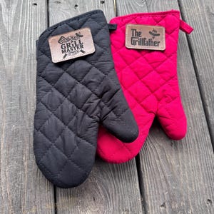 May include: Two quilted oven mitts, one black and one bright pink, rest on a wooden surface. Each mitt has a brown leather patch with white text. The black mitt reads "The Grill Master" and the pink mitt reads "The Grillfather".