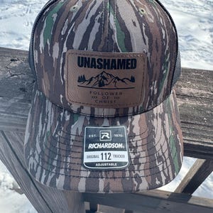 May include: A camouflage trucker hat with a brown leather patch that reads "Unashamed Follower of Christ" and a black mesh back. The hat is adjustable and has the Richardson logo and "Original 112 Trucker" printed on the back.