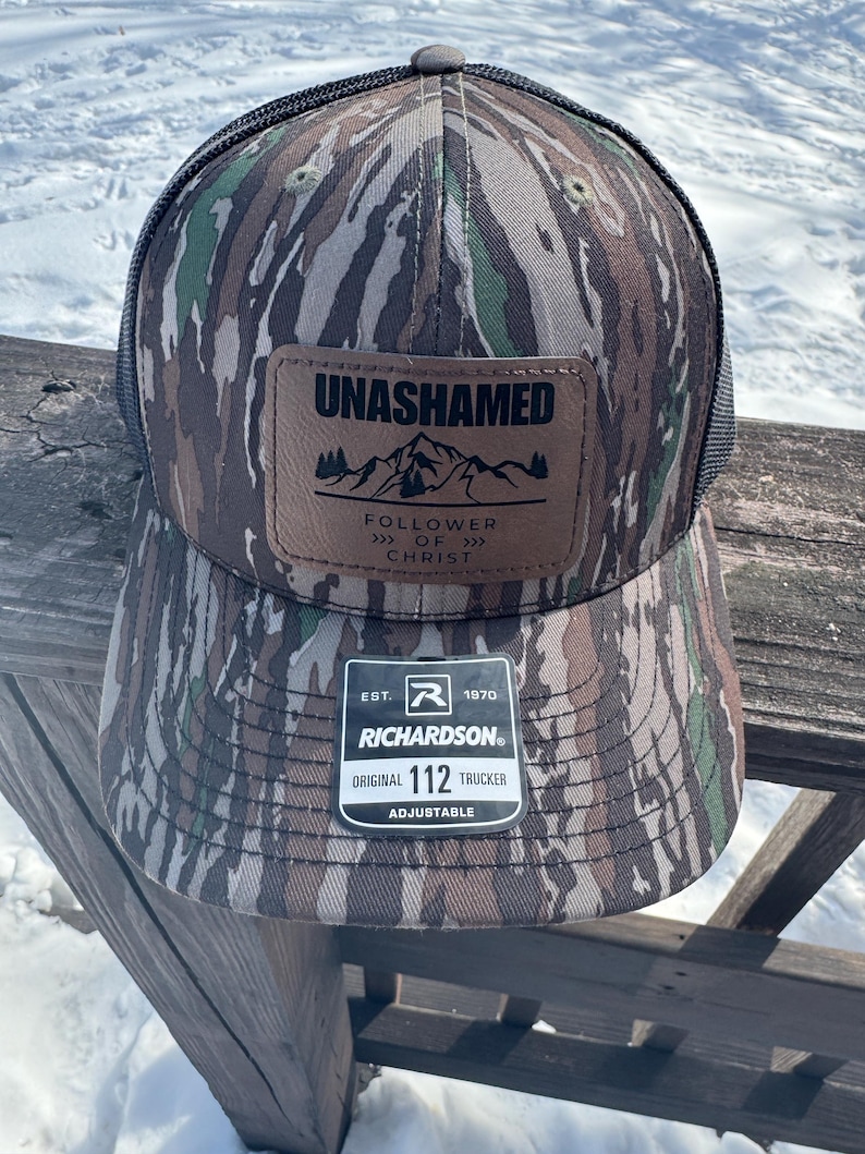 May include: A camouflage trucker hat with a brown leather patch that reads "Unashamed Follower of Christ" and a black mesh back. The hat is adjustable and has the Richardson logo and "Original 112 Trucker" printed on the back.