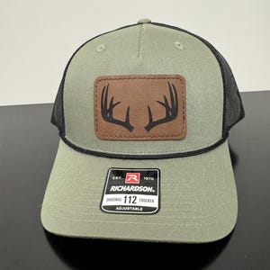 May include: A green and black trucker hat with a brown leather patch featuring a black antler design. The hat has a curved brim and an adjustable strap. The label reads "RICHARDSON 112 TRUCKER".