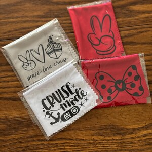 May include: Four different microfiber cleaning cloths with various designs. The cloths are packaged in clear plastic bags. The designs include a peace sign, a heart, a cruise ship, a Mickey Mouse hand, a bow, and the text "Cruise Mode On".