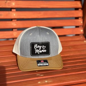 May include: A gray and white trucker hat with a brown brim. The hat has a black patch with the text "Boy Mama" in white with a heart symbol.