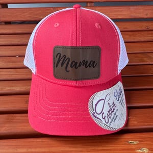 May include: A red and white trucker hat with a brown leather patch that says "Mama" on it. The hat has a mesh back and a concealed ponytail opening.