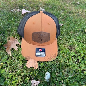 May include: A brown and black baseball cap with a black mesh back. The front of the cap has a brown leather patch with the letters "M.I." and a small pine tree graphic. The cap has a white label with the text "Richardson 115" on the side.