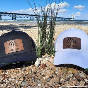 May include: Two black and white trucker hats with brown leather patches. The patches have the words "Mr. EST. 2024" and "Mrs. EST. 2024" on them.