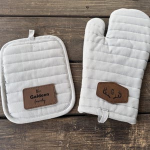 May include: A light gray quilted oven mitt and pot holder set. The pot holder has the text "The Galdeen Family" on a brown leather patch. The oven mitt has a brown leather patch with a deer antler design and the letter "G".
