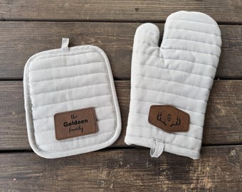 May include: A light gray quilted oven mitt and pot holder set. The pot holder has the text "The Galdeen Family" on a brown leather patch. The oven mitt has a brown leather patch with a deer antler design and the letter "G".
