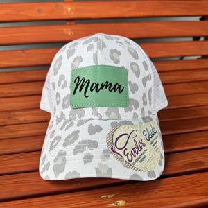 May include: White and gray leopard print trucker hat with a green patch that says "Mama" on it. The hat has a concealed ponytail opening and a small logo on the side that says "Evolve Eludes".