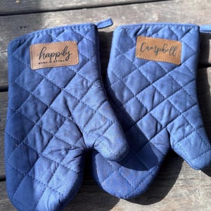 May include: Two navy blue quilted oven mitts with brown leatherette patches. One mitt reads "happily ever after" and the other reads "Campbell" in script. The mitts are made of a diamond-patterned fabric.