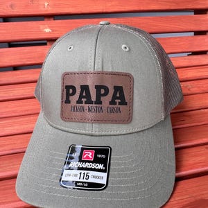 May include: A green trucker hat with a mesh back. The hat features a brown leather patch with the word "PAPA" in bold letters, and the names "JACKSON - WESTON - CARSON" below. A black and white tag is attached to the hat.