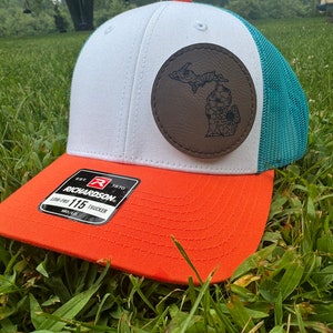 May include: A trucker hat with a white front panel, orange brim, and teal mesh back. A brown leather patch features a floral design of the state of Michigan. The hat has a Richardson tag.