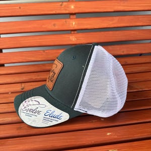 Pu&ograve; includere: Un cappello da camionista verde scuro con una rete bianca sul retro. Il cappello ha una toppa in pelle marrone con il testo "Stay Wild" su di essa. Il cappello ha anche un'etichetta bianca con il testo "Evolve" e "Elude" su di essa. L'etichetta dice anche "Apertura per coda di cavallo nascosta" e "Fascia anti-sudore resistente al trucco".