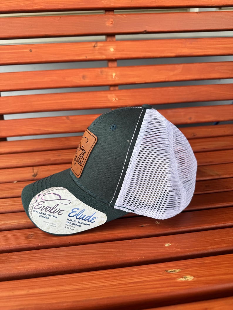 Pu&ograve; includere: Un cappello da camionista verde scuro con una rete bianca sul retro. Il cappello ha una toppa in pelle marrone con il testo "Stay Wild" su di essa. Il cappello ha anche un'etichetta bianca con il testo "Evolve" e "Elude" su di essa. L'etichetta dice anche "Apertura per coda di cavallo nascosta" e "Fascia anti-sudore resistente al trucco".